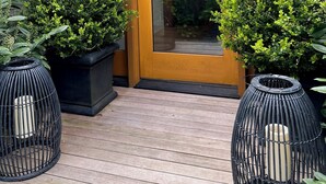 A wooden door with a glass panel, flanked by potted plants and two black wicker chairs on a wooden deck.