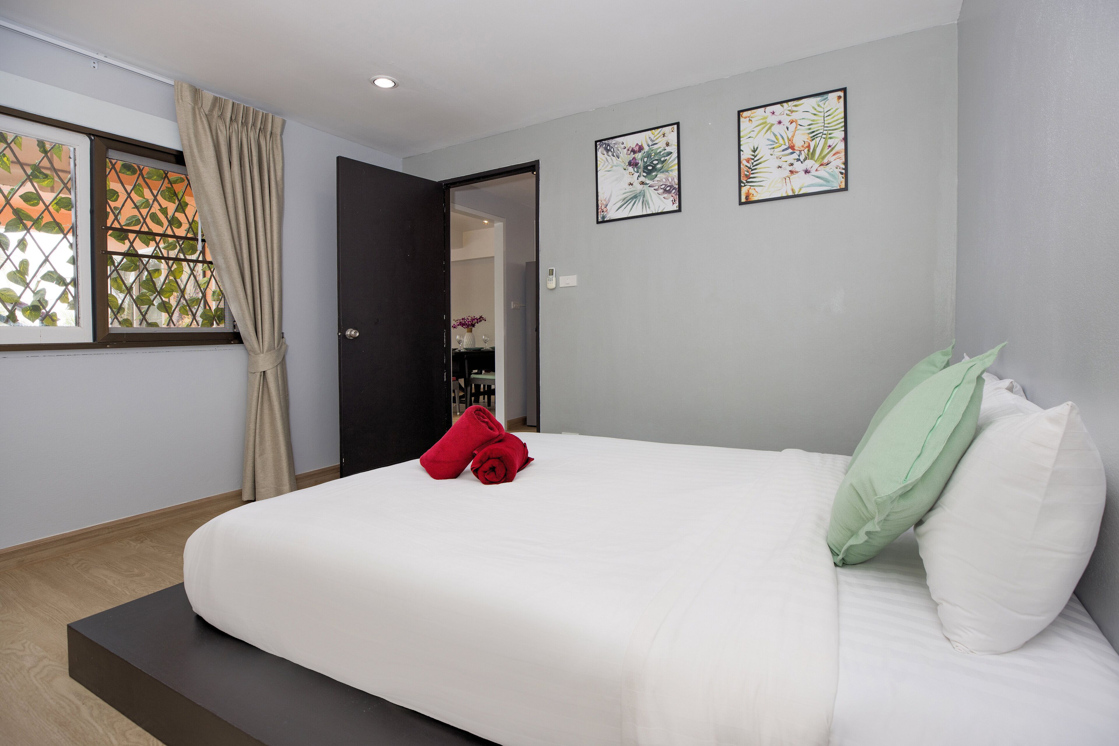 Una cama bien hecha con ropa blanca y una almohada verde, una toalla roja y una cortina color beige en una ventana con diseño de celosía.