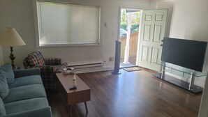 A living room with a sofa, a coffee table, a TV on a glass stand, a heater, and a door leading outside.