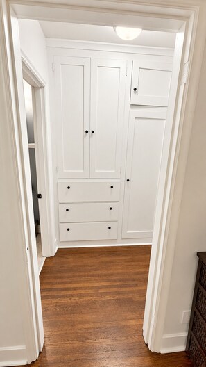 A walk-in closet with white doors and drawers, wooden flooring, and a ceiling light.