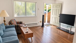 A living room with a sofa, a coffee table, a TV on a glass stand, a heater, and a door leading outside.