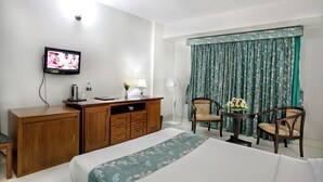 A hotel room with a bed, a wooden desk, a television, a desk lamp, a fan, two chairs, and a window with curtains. - Hotel Purbani International Limited (Dhaka)