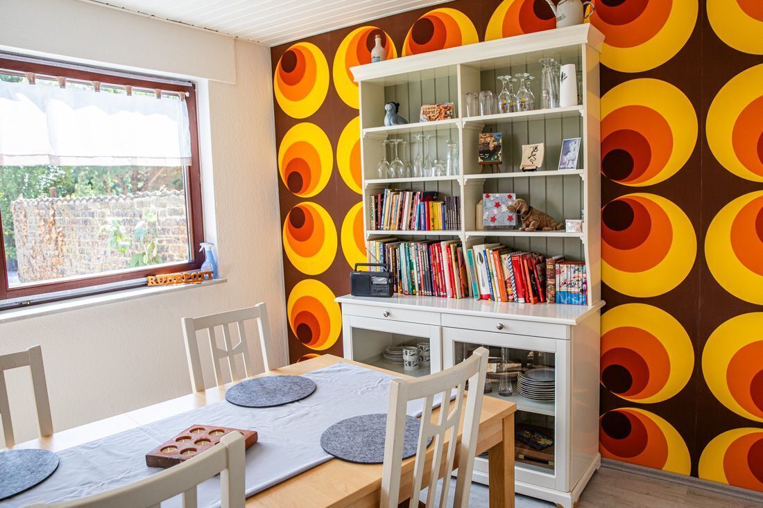 Ein Essbereich mit Tisch für zwei Personen, einer Bücherwand mit Büchern und Dekoration, sowie ein Fenster mit Blick auf einen Garten.