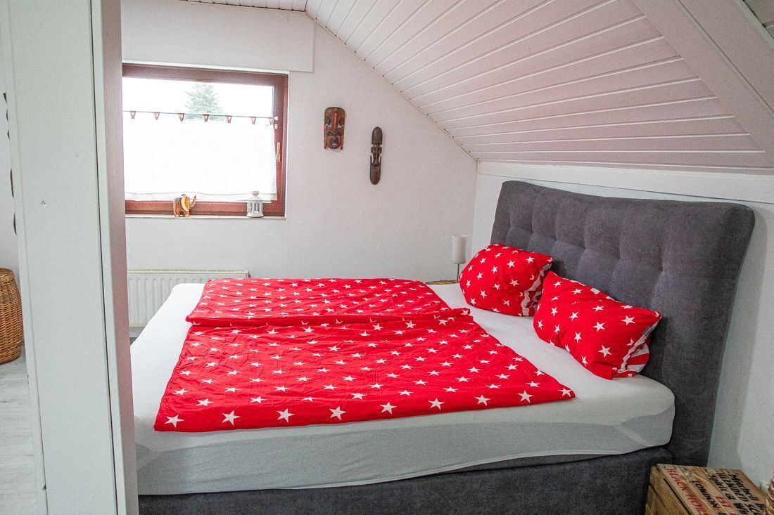 Une chambre avec un plafond en pente, un lit doté d’une tête de lit grise et une fenêtre donnant sur l’extérieur.