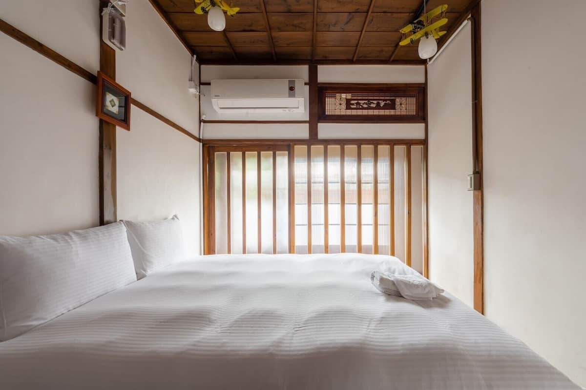 A bedroom with a bed, wooden beams, a window with vertical blinds, and an air conditioning unit.
