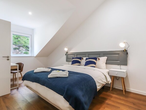 A bedroom with a sloped ceiling, a bed with a blue and white bedspread, two bedside tables with lamps, and a window with a view of greenery.