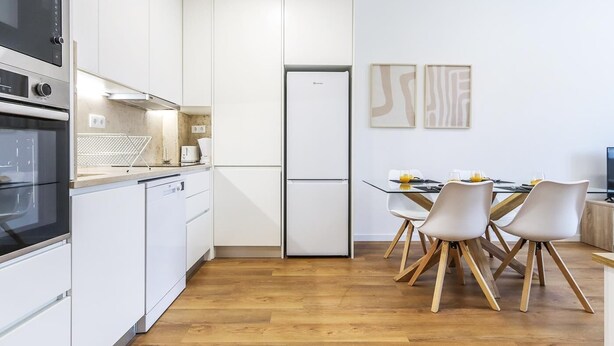 Et moderne køkken med hvide skabe, en indbygget ovn og et køleskab. Der er et spiseområde med et bord og stole.