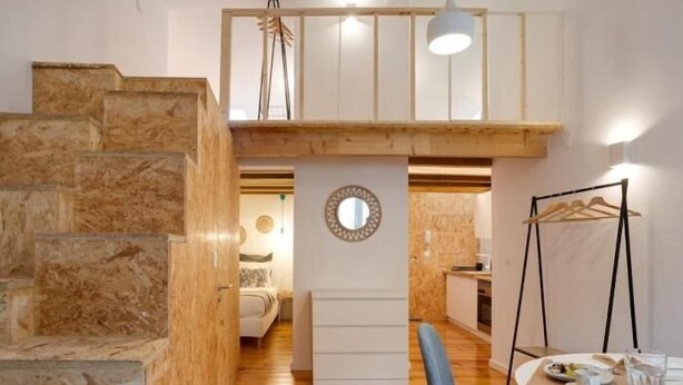 A modern interior with a wooden staircase, a loft bedroom with a bed, a dining area with a table and chairs, and a kitchen area.