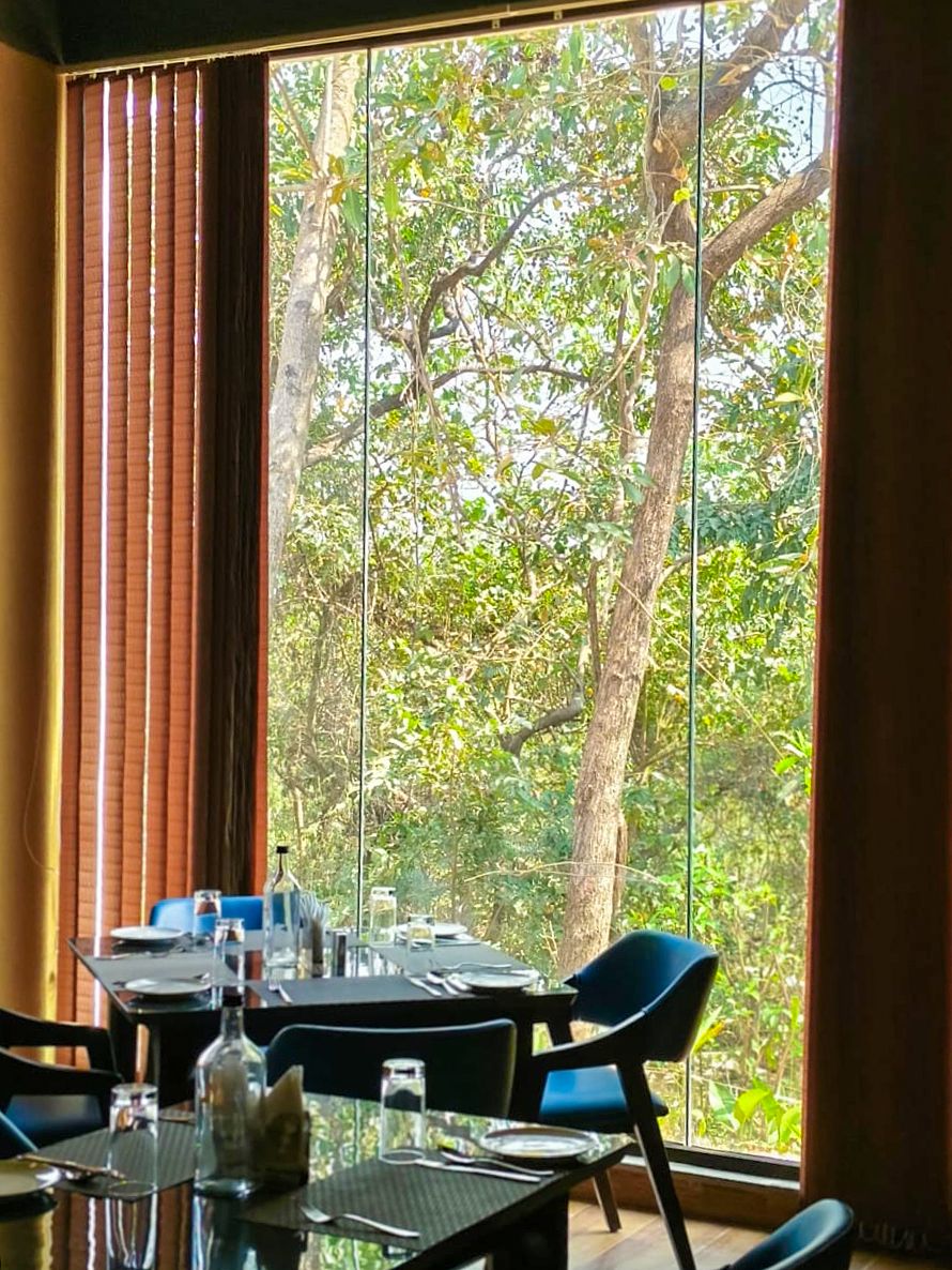 A dining area with a table set for two, large windows with a view of trees, and brown curtains.