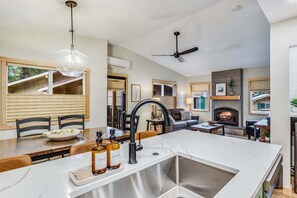 A modern kitchen with a large island, a dining area with wooden chairs, a fireplace, and a ceiling fan.