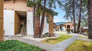 Un bâtiment moderne doté d’une façade en bois, d’un escalier apparent menant à l’entrée, et entouré d’un paysage verdoyant avec des arbres et un sentier.