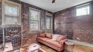 Una habitación con paredes de ladrillo, un sofá rosa, una mesita, una silla y un ventilador de techo.