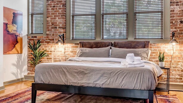 Un dormitorio moderno con pared de ladrillo, ventanas grandes con persianas, una cama con colcha gris, una mesita de noche pequeña con lámpara y una planta en maceta.