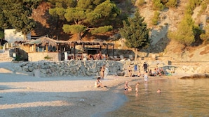 A beach with a stone wall, a thatched-roof structure, and people in the water. - Apartment Tafra 2 (Omis)
