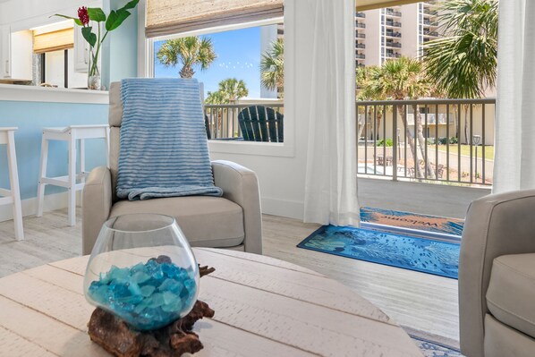 A balcony with a view of palm trees and a pool, a chair with a striped cushion, and a glass bowl with blue crystals on a wooden table.