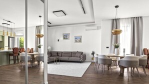 Een moderne woonkamer met een grijze bank, een eettafel en stoelen. Er is een grote spiegel die de kamer weerspiegelt, een wit plafond en een raam met gordijnen.