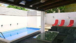 An outdoor pool area with a glass table and red chairs, surrounded by a white wall and covered with a striped awning. - Holiday Home Mumy (Dobrinj)