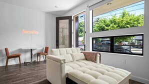 Un salon moderne avec un canapé, deux fauteuils, un panneau lumineux au néon et une grande fenêtre donnant sur des arbres et des voitures garées.