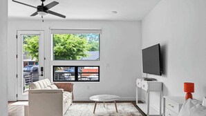 Un salon moderne avec un canapé blanc, une petite table ronde, un téléviseur posé sur un meuble et une vue sur l’extérieur grâce à une grande fenêtre.