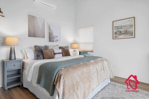 Un dormitorio con cama, dos mesitas de noche, una mesilla, decoración en la pared y una ventana con persianas.