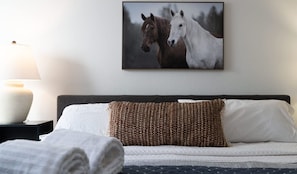 A neatly made bed with a patterned pillow, a framed picture of horses on the wall, and a bedside lamp.
