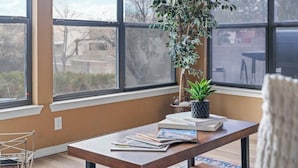 Ein Zimmer mit großen Fenstern, einem Holztisch mit Büchern und einer Pflanze, und Blick auf Bäume und Gebäude.