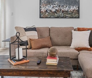 Una sala de estar con sofá, mesa de centro, control remoto, libros, una vela y un objeto decorativo.