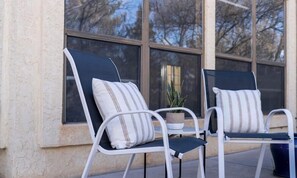 Two white metal chairs with striped cushions, a small potted plant, and a window with a view of trees.