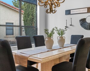 A dining area with a wooden table, black chairs, two white vases with plants, a chandelier, and a wall with a sign saying 'COME IN AND Stay awhile'.