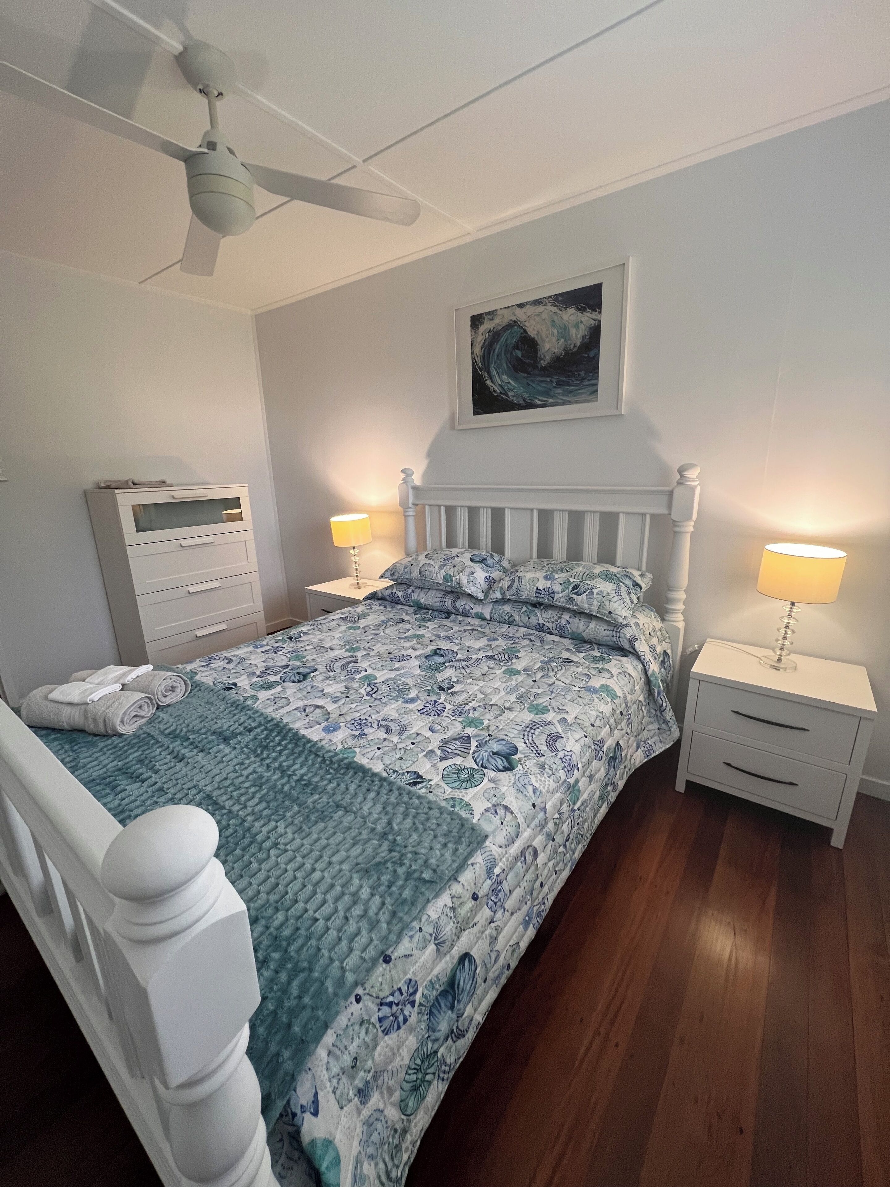 Un dormitorio con cama, mesitas de noche, ventilador de techo, calefacción en la pared y un cuadro.