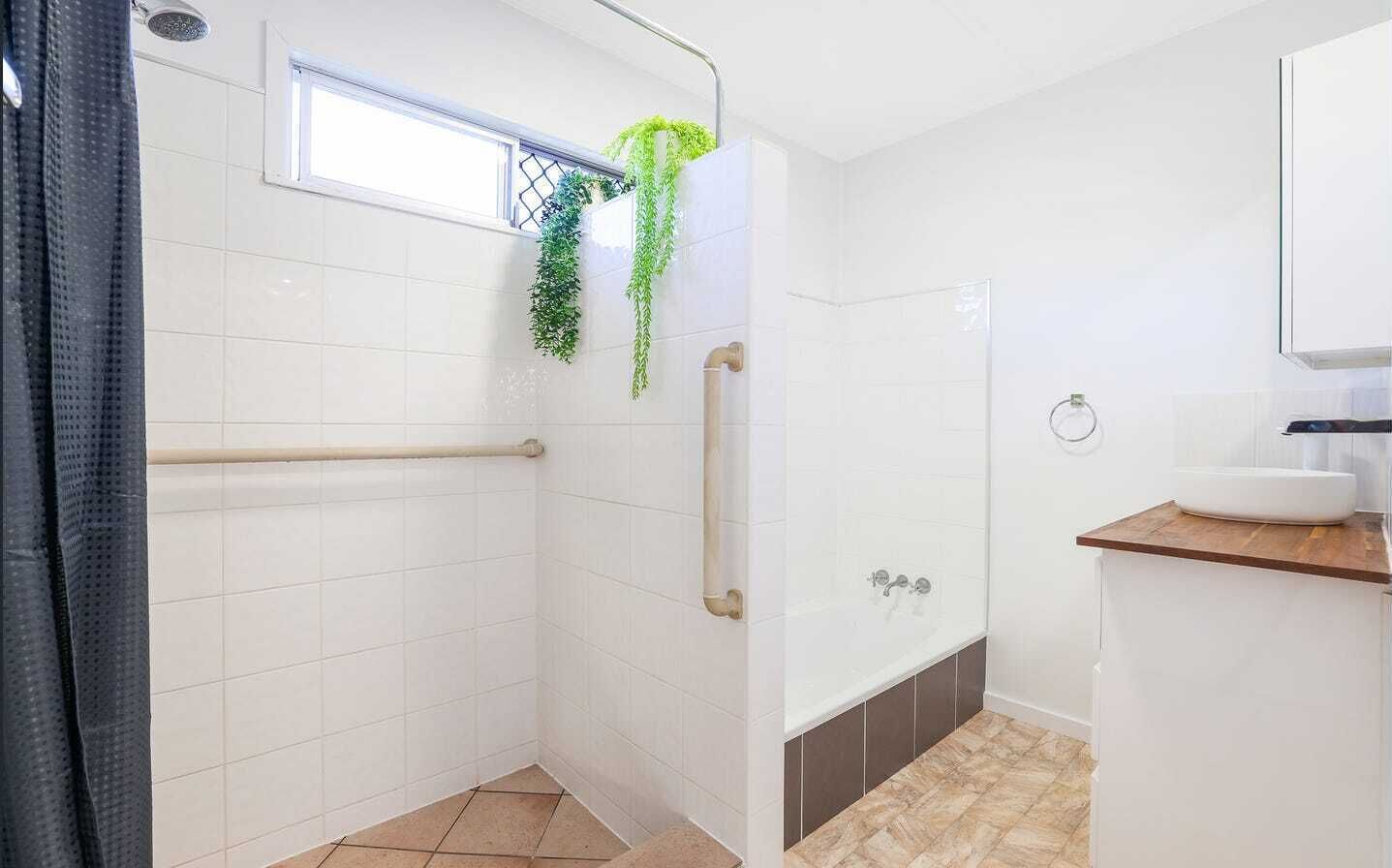 Un baño moderno con azulejos blancos, cabina de ducha de vidrio, tocador de madera y ventana sobre el lavabo.