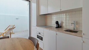 A modern kitchen with white cabinets, a sink, and a coffee machine. There is a dining table with chairs and a window overlooking a white wall. - Simple Life I, Setúbal (Setúbal)