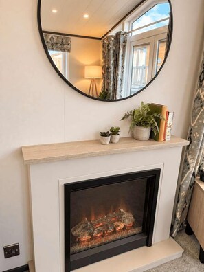 A fireplace with a mirror above it, a shelf with plants and books, and a window with curtains.