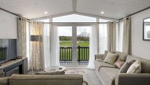 Amplia sala de estar con televisor grande, sofá y mesa de centro. Hay un balcón con vistas a un paisaje verde.