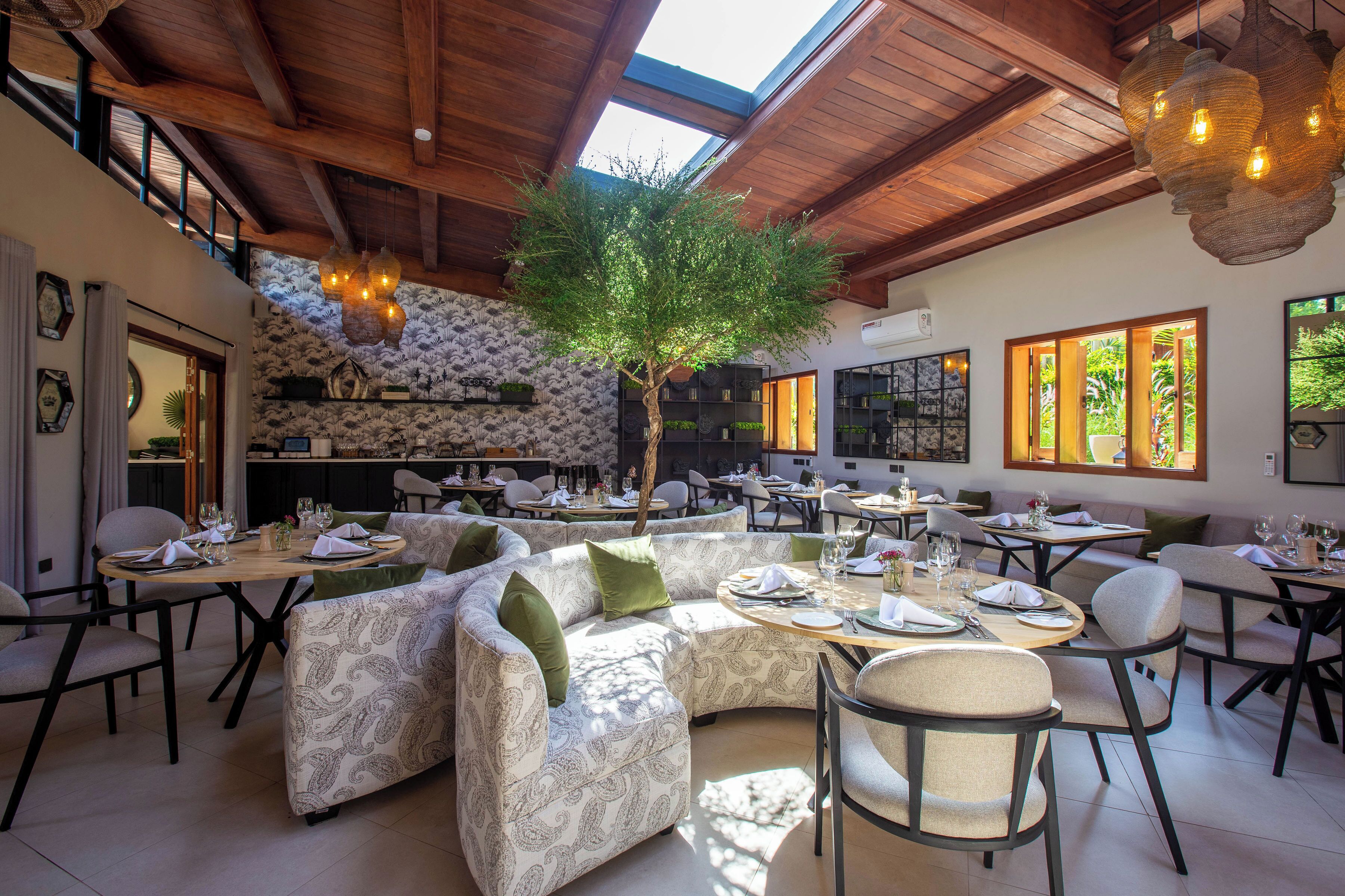 A spacious dining area with a high ceiling, a central tree, and multiple dining tables set for guests.