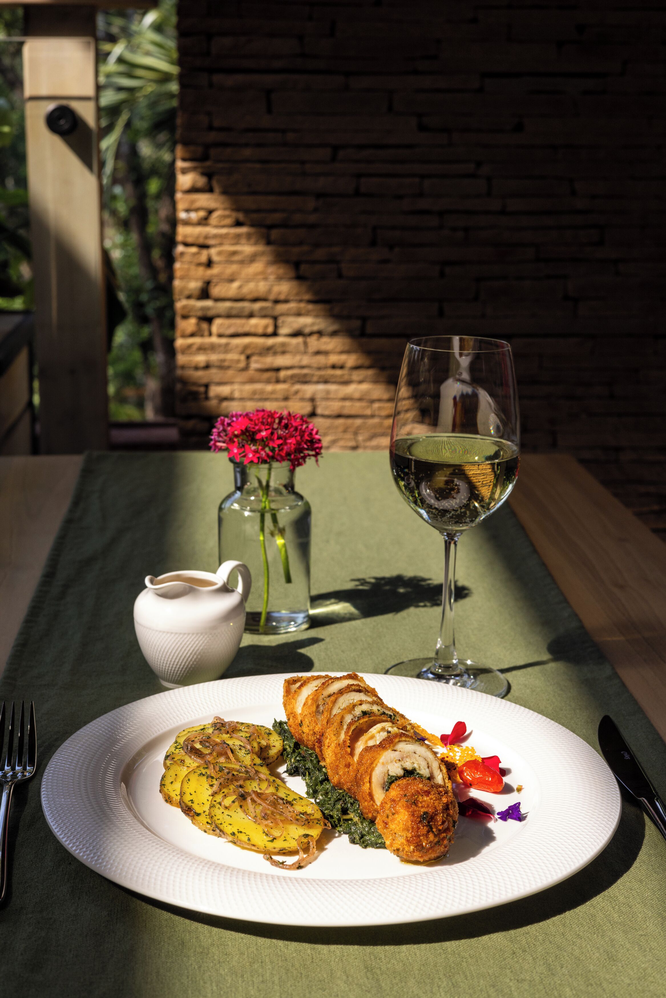 Un plato con comida, una copa de vino blanco, un florero con flores y una jarra pequeña sobre una mesa.