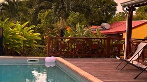 A pool area with a wooden deck, surrounded by lush greenery and a building with a red roof. - Bungalows in Guadeloupe ATOUMO (Petit-Canal)