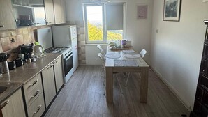 A compact kitchen with a dining area, a refrigerator, a stove, and a window with a view of trees. - Plesivica Hillside Holiday Home With Hot -tub (Jastrebarsko)