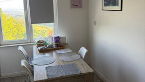 A dining area with a table set for two, a window with a view of a yellow landscape, and a framed picture on the wall. - Plesivica Hillside Holiday Home With Hot -tub (Jastrebarsko)