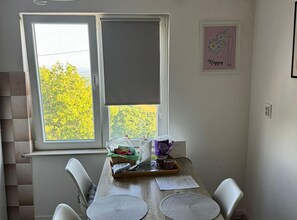 A dining area with a table set for two, a window with a view of trees, and a framed picture on the wall.