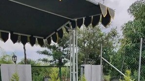 A white metal swing bench under a black canopy with a decorative edge, set in a grassy outdoor area with trees and a stone wall in the background.