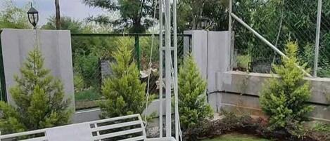 Una panca a dondolo in metallo bianco sotto un gazebo nero con bordo decorativo, immersa in un'area verde con alberi e un muro in pietra sullo sfondo.