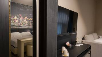 A hotel room with a bed, a bedside table with a cup and a remote control, a large flat-screen TV mounted on the wall, and a window with curtains.