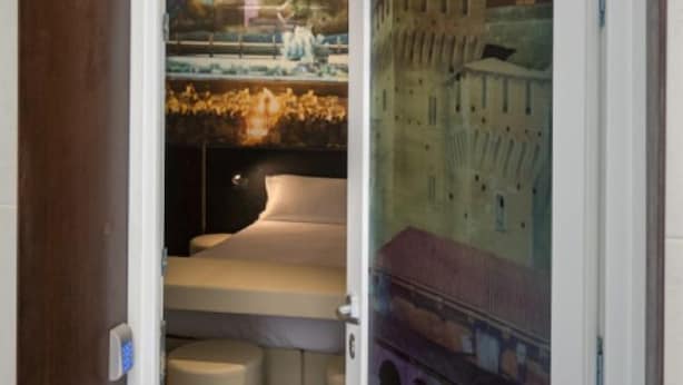 Une chambre d’hôtel avec un lit, une petite table et une vue sur un bâtiment historique, visible à travers une porte vitrée.