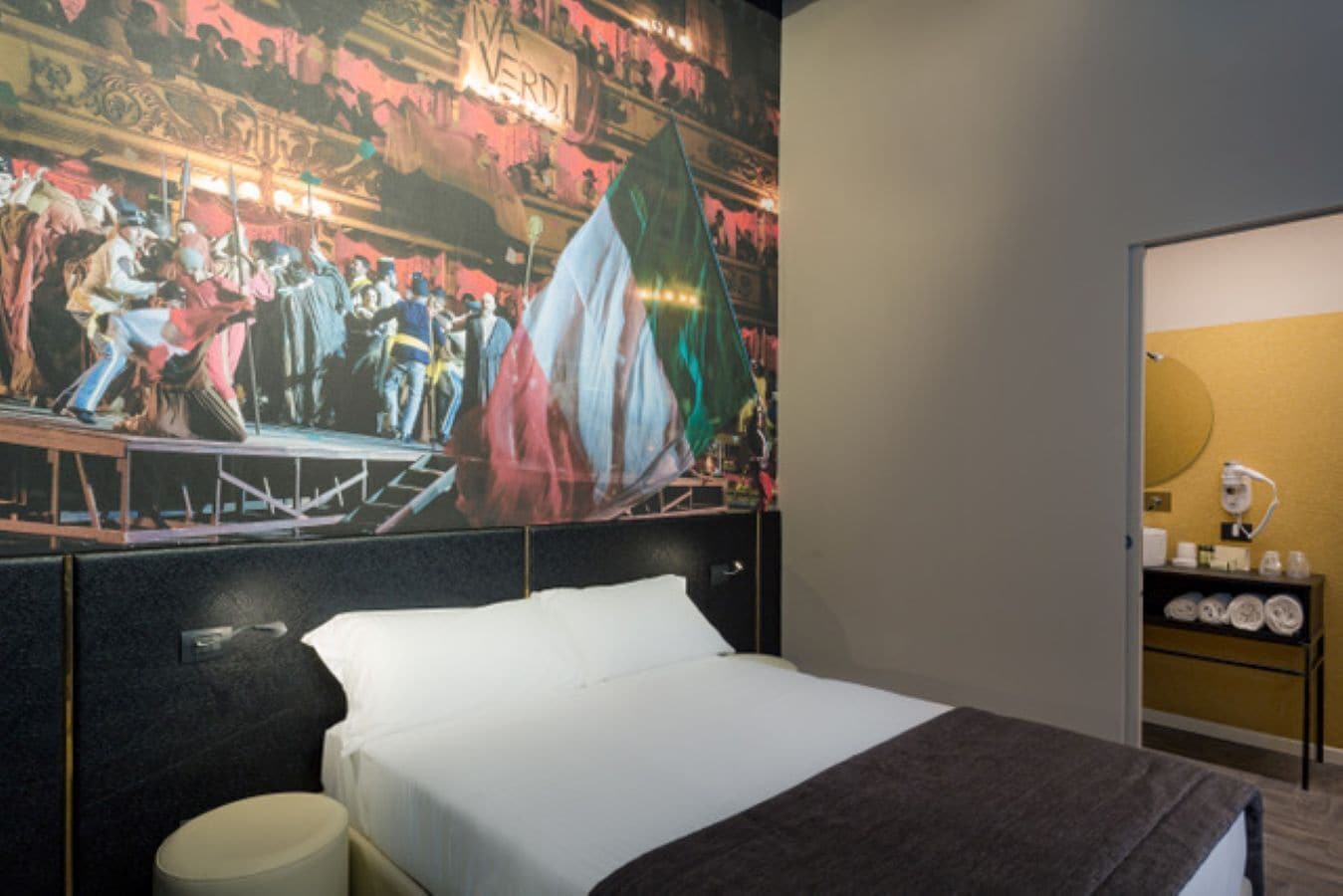 A hotel room with a bed, a nightstand, a wall-mounted shelf with toiletries, and a large mural depicting a historical scene.