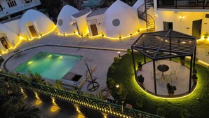 A pool area with a gazebo, surrounded by uniquely shaped buildings with circular windows and warm lighting. - Shridhar Reesort (Rishikesh)