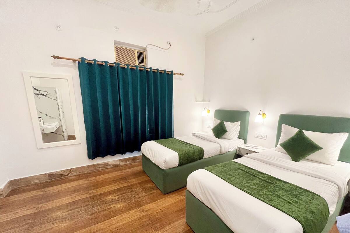 A hotel room with two beds, a wooden floor, a window with curtains, and a small air conditioning unit.