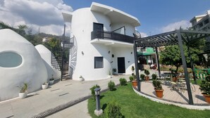A modern building with a unique architectural design, featuring a curved facade, a balcony with black railings, and a covered outdoor seating area with tables and chairs. - Shridhar Reesort (Rishikesh)
