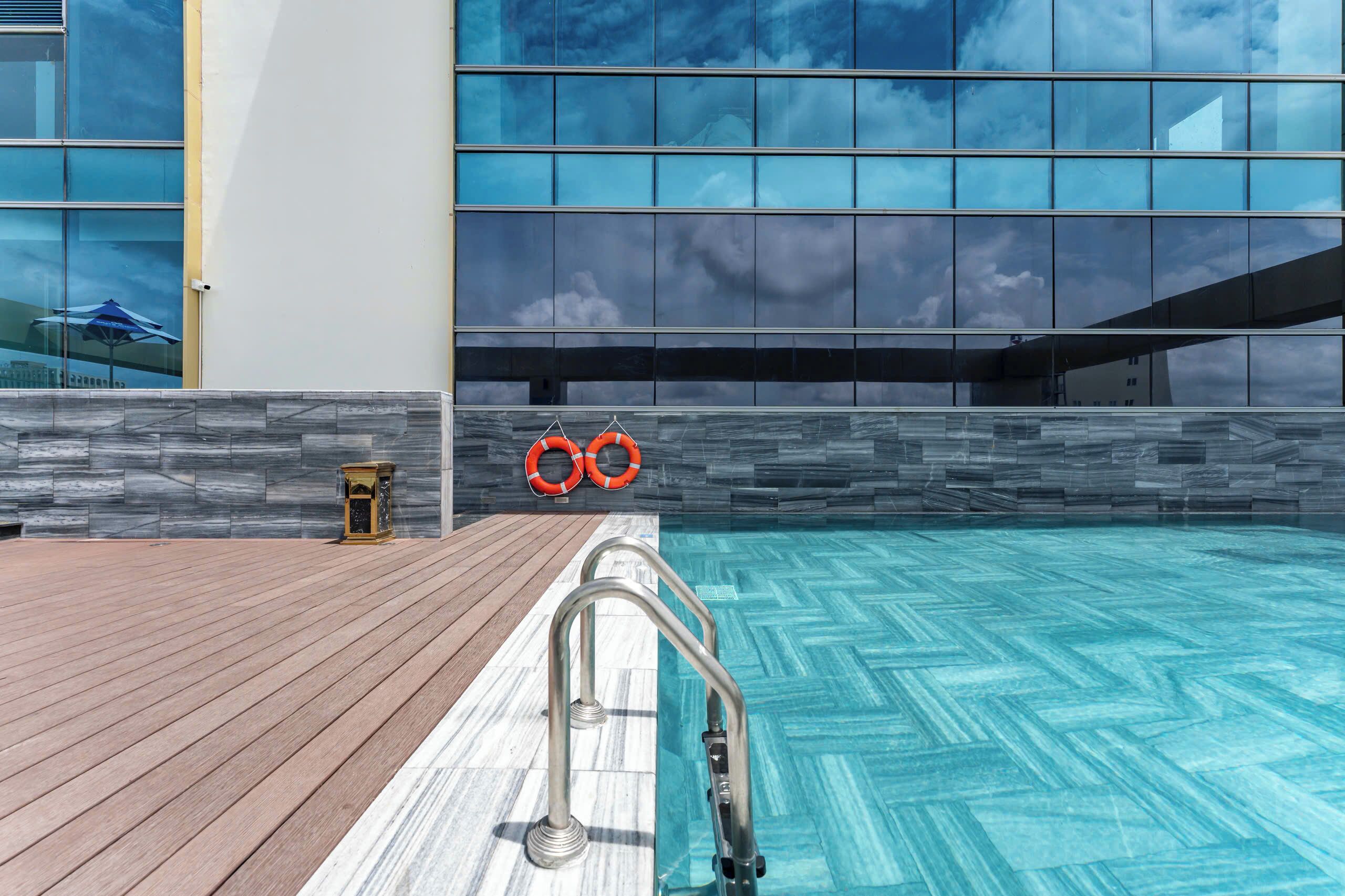 Zona de piscina con escalera metálica, cubierta de madera, pared de piedra y un edificio de cristal al fondo.