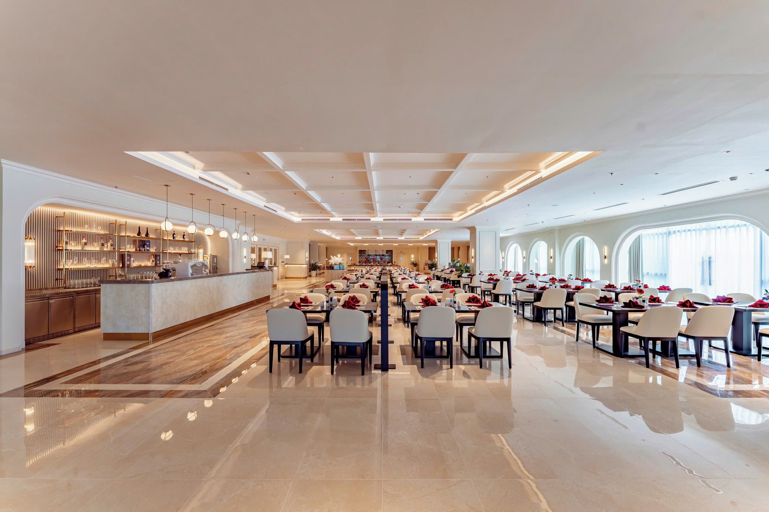 A spacious hotel lobby with a long reception desk, multiple dining areas, and large windows.